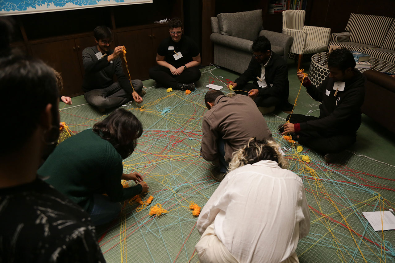 8 players sit on a large net on the ground. Most of them have colorful balls of yard in their hands and are busy with weaving it into the net right where they sit. Some cozy couches are in the background.