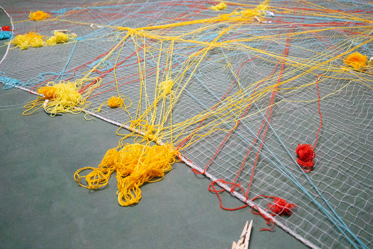 A bright net is photographed from the side, with yellow, red, and blue yarn woven in and out of the grid. The different strings form nodes in positions where many strings meet. Close to the camera, there is a big heap of yellow yarn on the floor with many strings reaching back into the net.