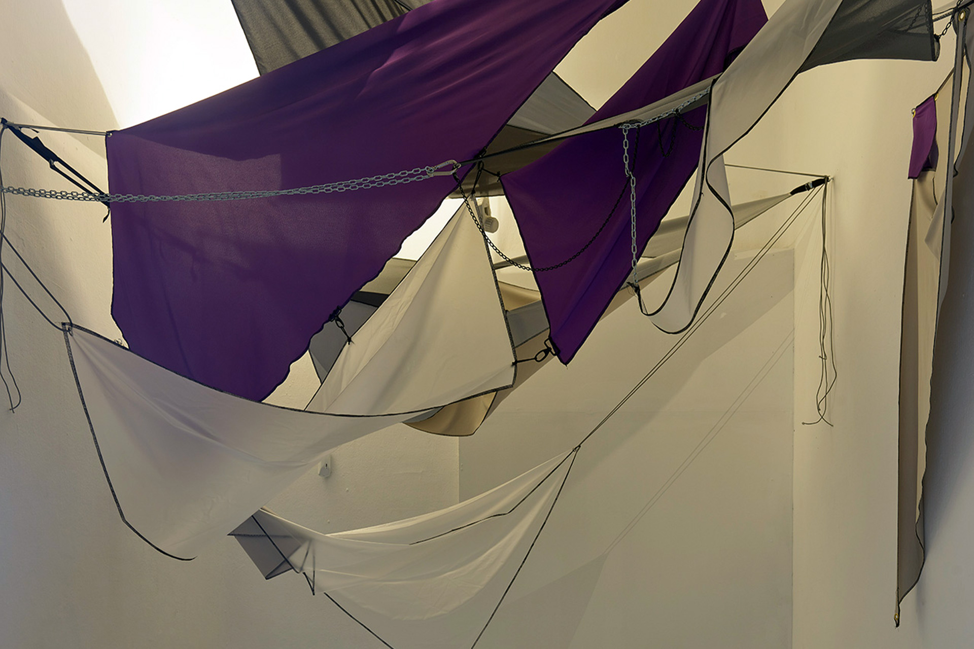 Room with geometric fabric art installation in shades of purple, gray, and beige suspended from the ceiling. The fabric pieces each stand for a star costellation and are held together and hug with various hooks, chains and zippers. Two round gray cushions with shiny silver tops are on the glossy floor below, with star constellations charts atop.
