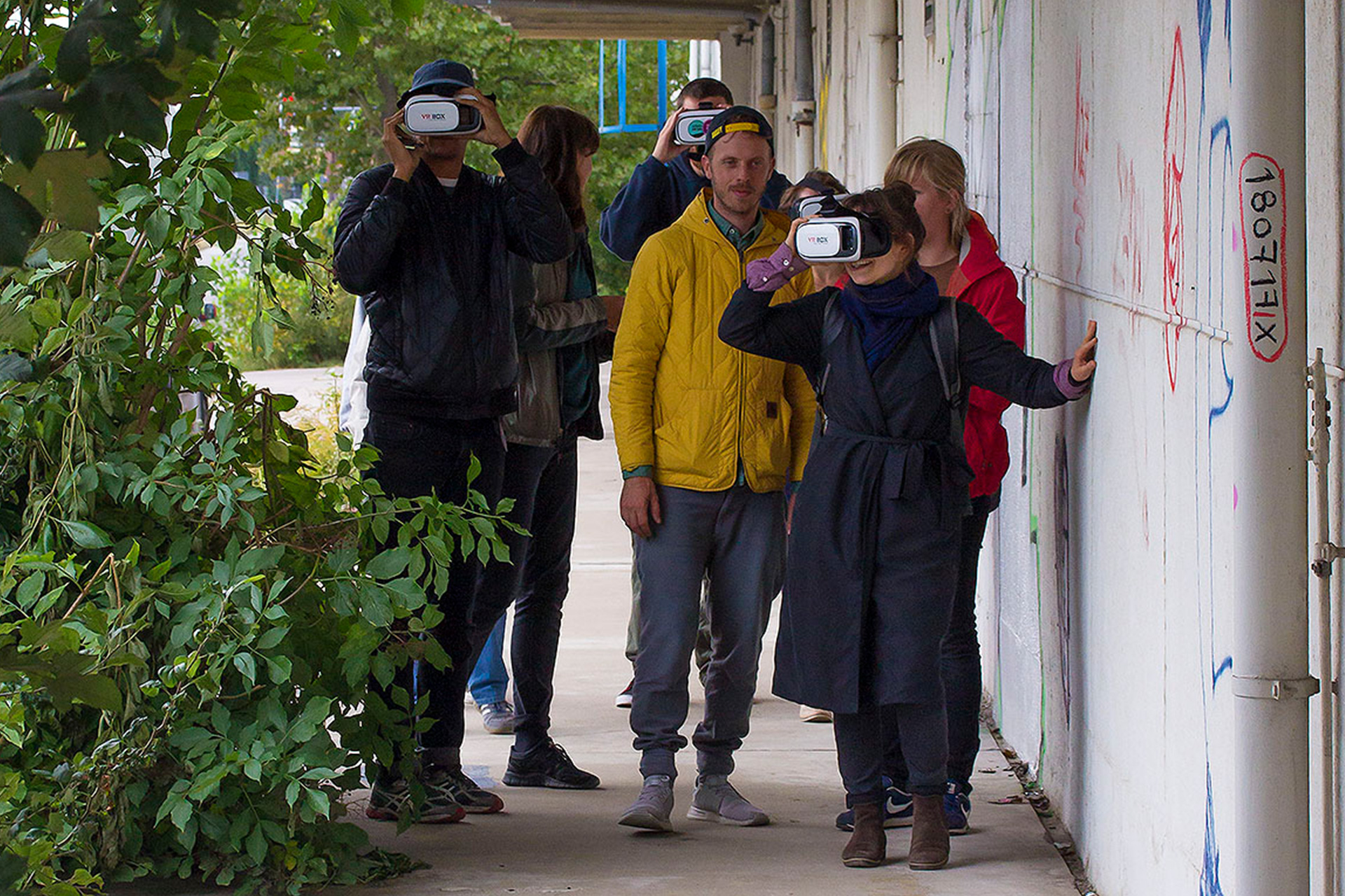 On an Augmented Reality Walk, three out of six participants are wearing white AR headsets. The person in the front is touching the headset with their hand, and using the other hand to touch the concrete wall next to them.