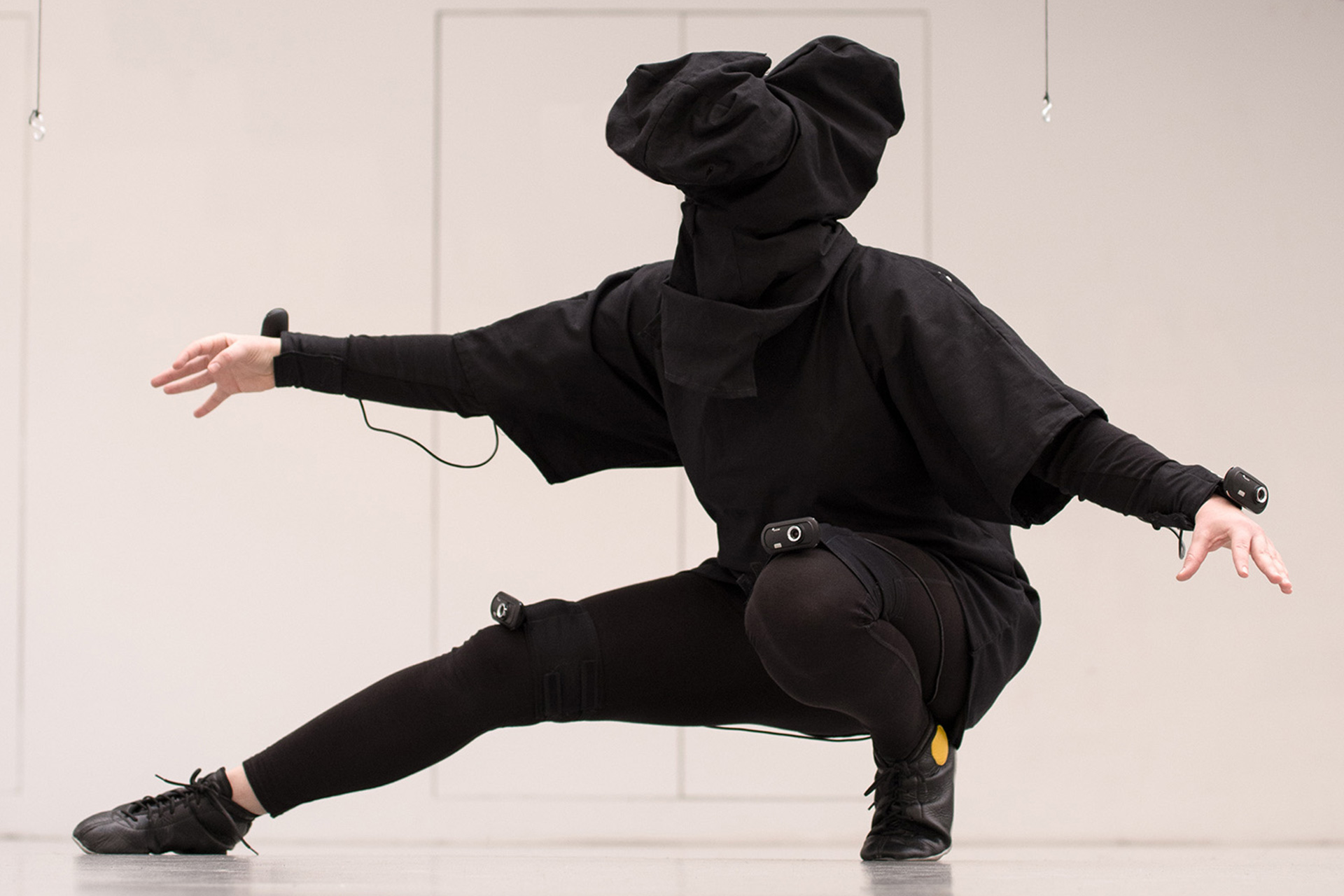 A person is wearing a dark, self-made Virtual Reality Suit that consists of a hood for the head in which a VR headset is hidden, a large shirt that has webcams attached to it, and leggins. They crouch down, with one leg extended and one leg holding their weight. Their arms are reaching towards the sides of the image. Webcams are attached to their wrists, and to both knees.