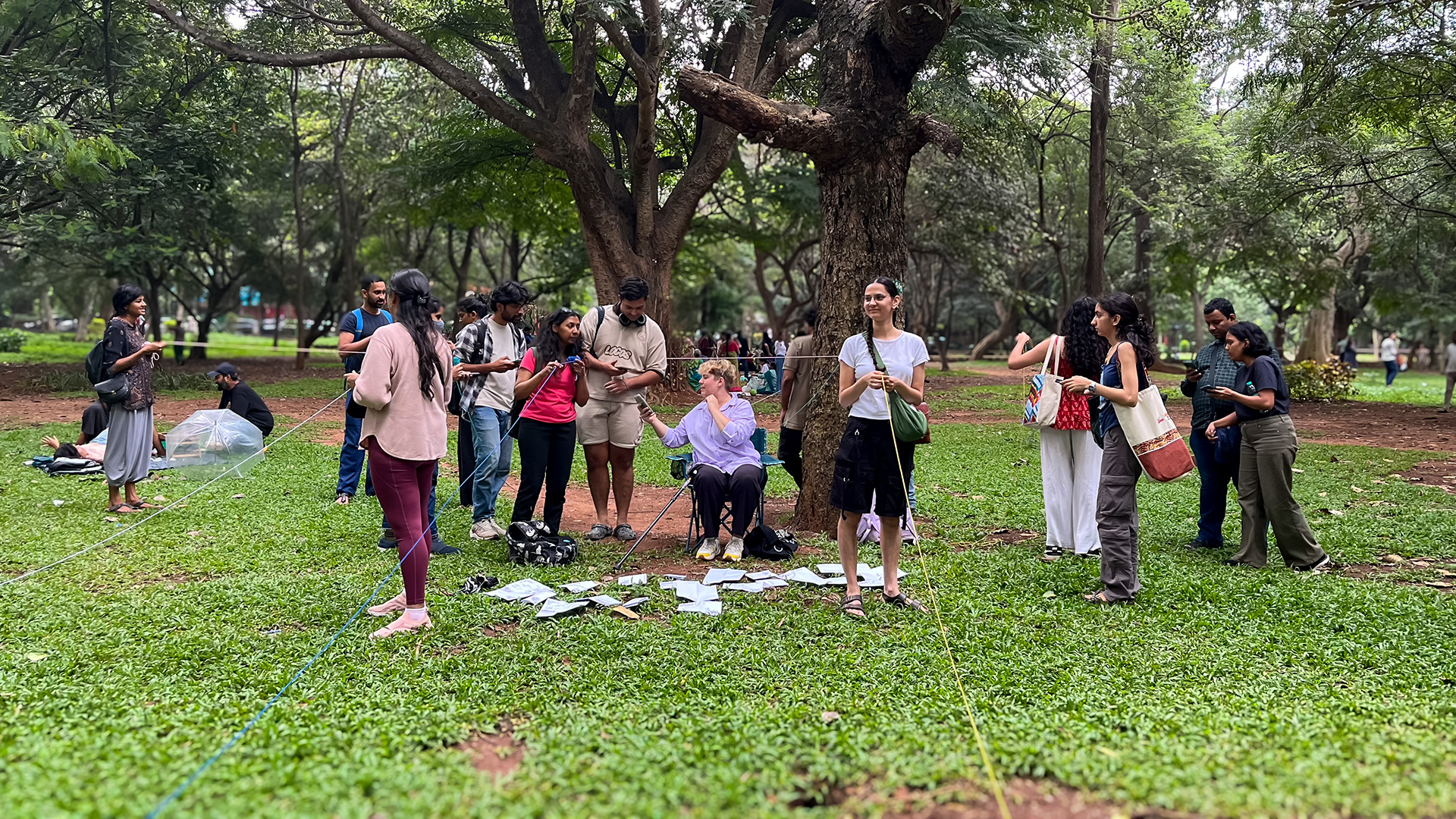 A group of people gathered on a lush green lawn in a park, surrounded by tall trees, are using strings to measure the dimensions of a hotspot. Some are standing while others sit. Papers are scattered on the grass. The setting is relaxed, with individuals engaging in conversation and the workshop activities.