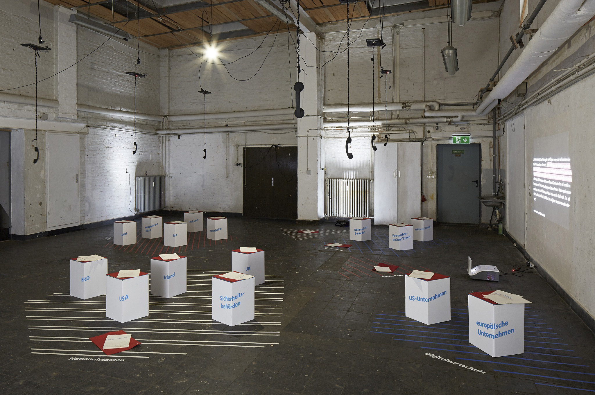 In a large industrial exhibition space, phones are hanging from the ceiling and dangling above low stools made from cardboard. The stools are arranged in groups atop grids made from tape on the floor. The group in the front right has the term Digitalwirtschaft printed on the floor, and stools that have europäische Unternehmen and US Unternehmen written on them. On wall behind these stools, a video is projected onto the wall.