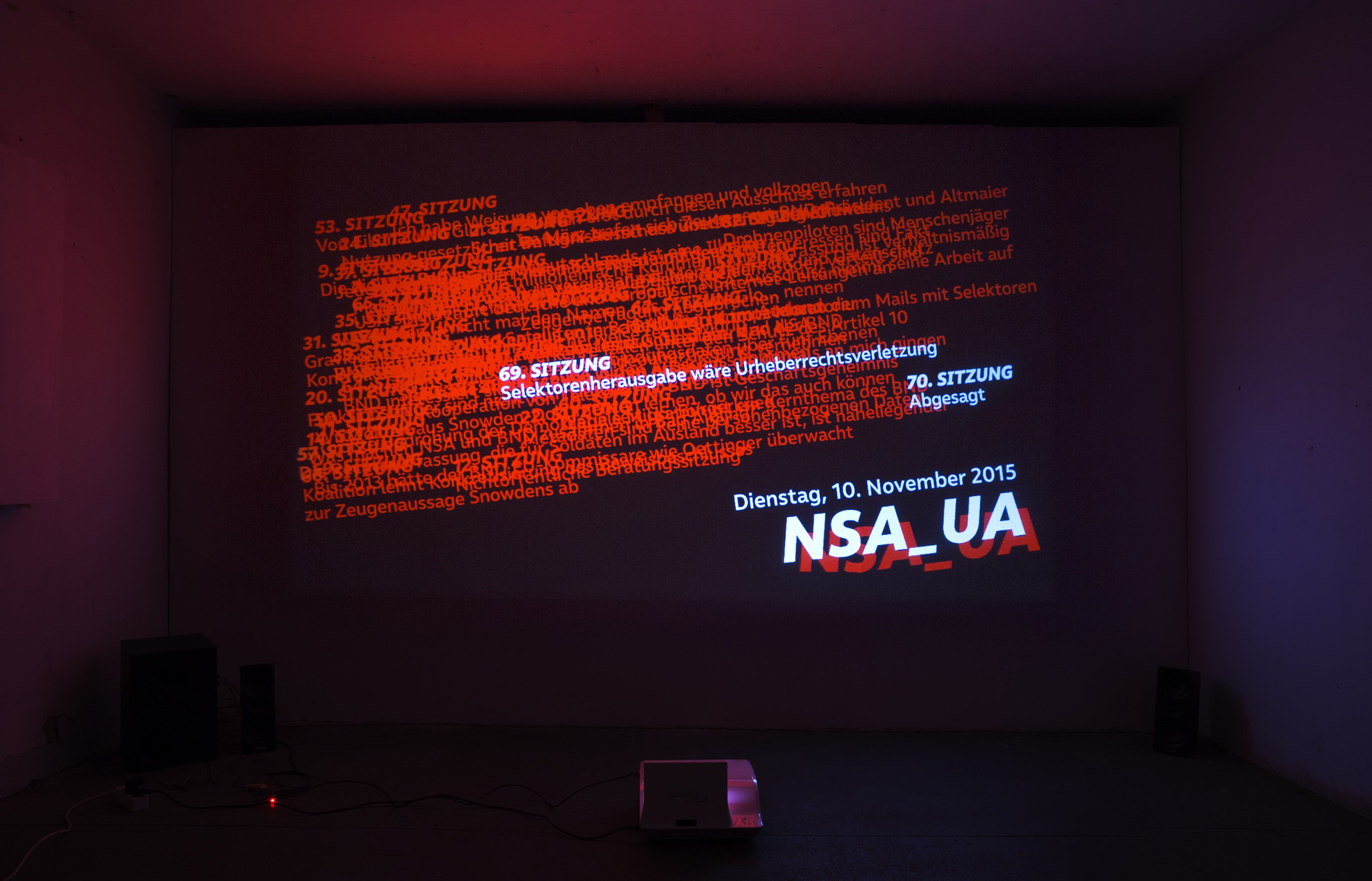 A video is projected onto a wall in a small, dark room. In the video, a large font reads a date: Tuesday, 10th of November 2015, above the letters NSA_UA. Above, the 69th session of the NSA_UA is highlighted, and many unreadable words are layered atop of each other.