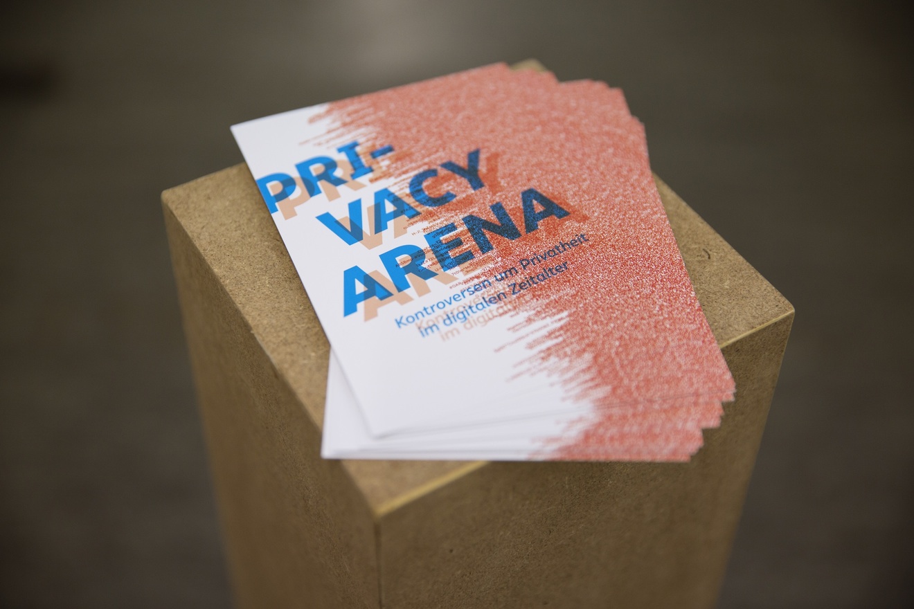 A few catalogues are on a pedestal so that visitors can pick them up. The cover of the catalogues has Privacy Arena written across it in blue, with a red pattern in the background.