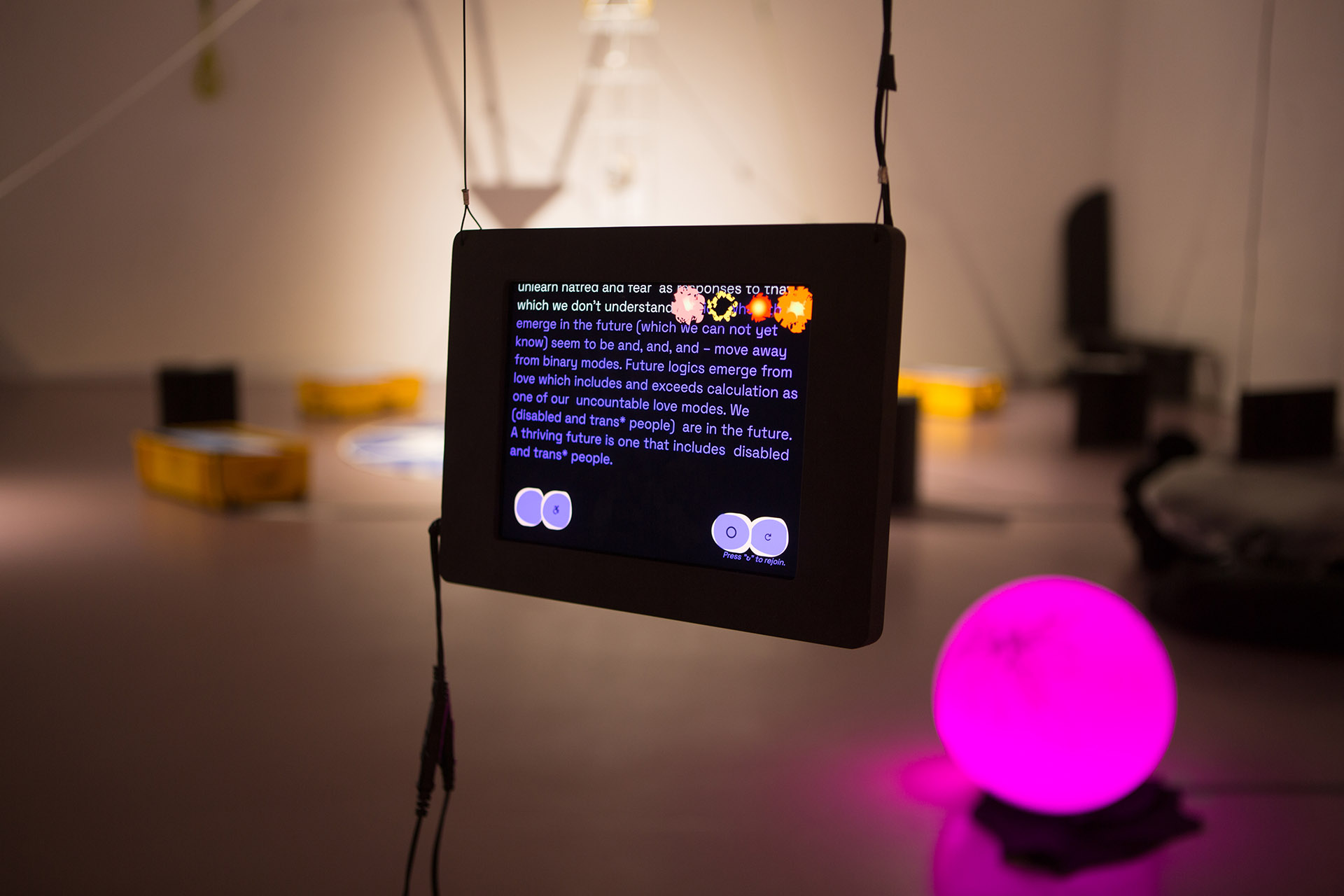 As part of the installation, a tablet hangs from the ceiling with the website artwork open. On it, sentences in violet color read: 'Future logics emerge from love which includes and exceeds calculation as one of our uncountable love modes. We (disabled and trans* people) are in the future. A thriving future is one that includes disabled and trans* people.' The background behind the tablet is blurry, and on the right side, a pink glass lampshade can be made out.