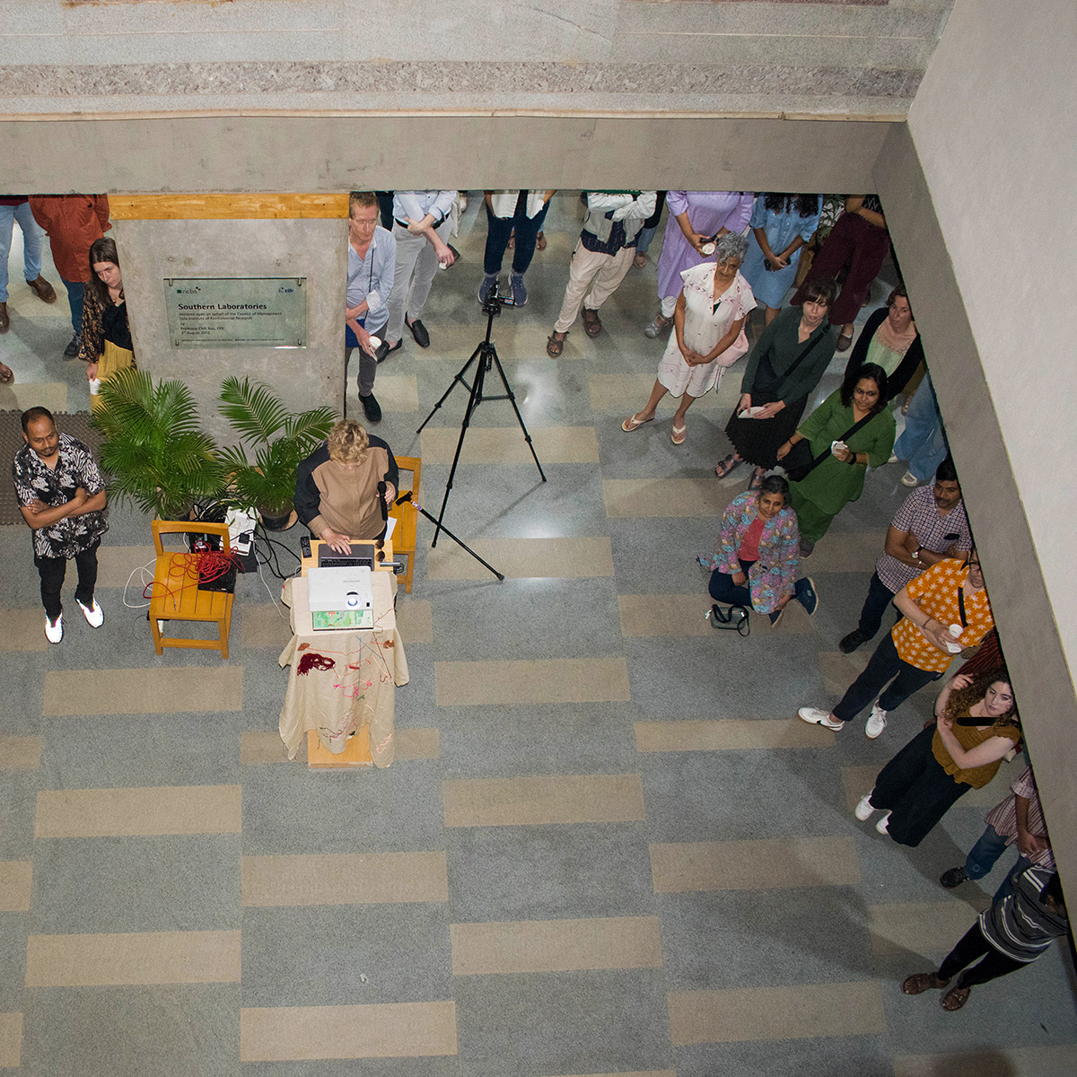 Overhead view of the opening of the art exhibition Sticky Networks with Iz, the artist, at a podium set up with equipment. The audience stands in a spacious hall with checkered flooring. People observe attentively.