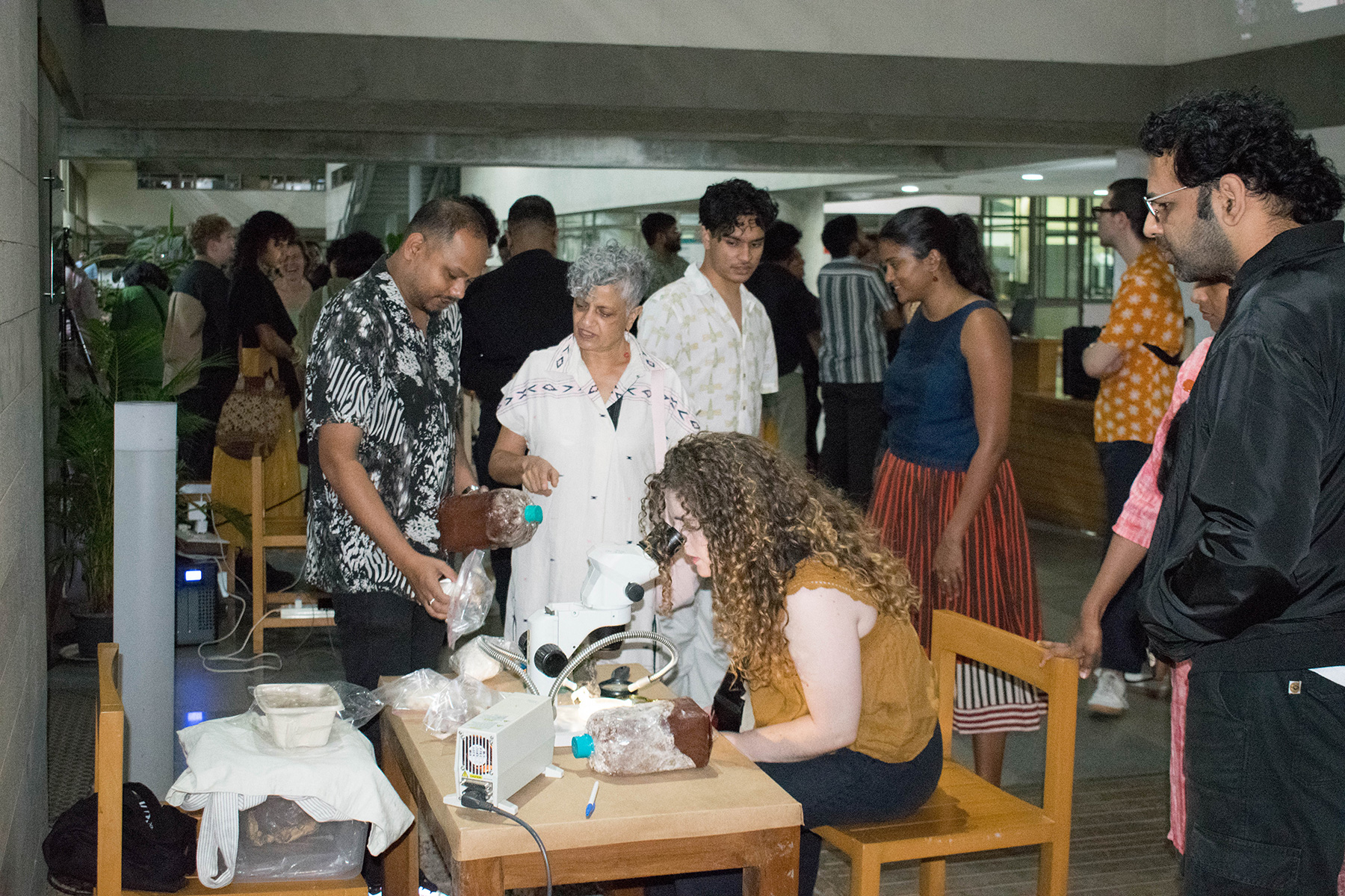 During the exhibition opening, a group of people stands around a woman using a microscope at a wooden table where Biplab Mahato introduces visitors to the world of mycelium. Various materials are scattered on the table: Spore bags, Petri dishes, and boxes with fungi samples. The background features tall windows and concrete elements.