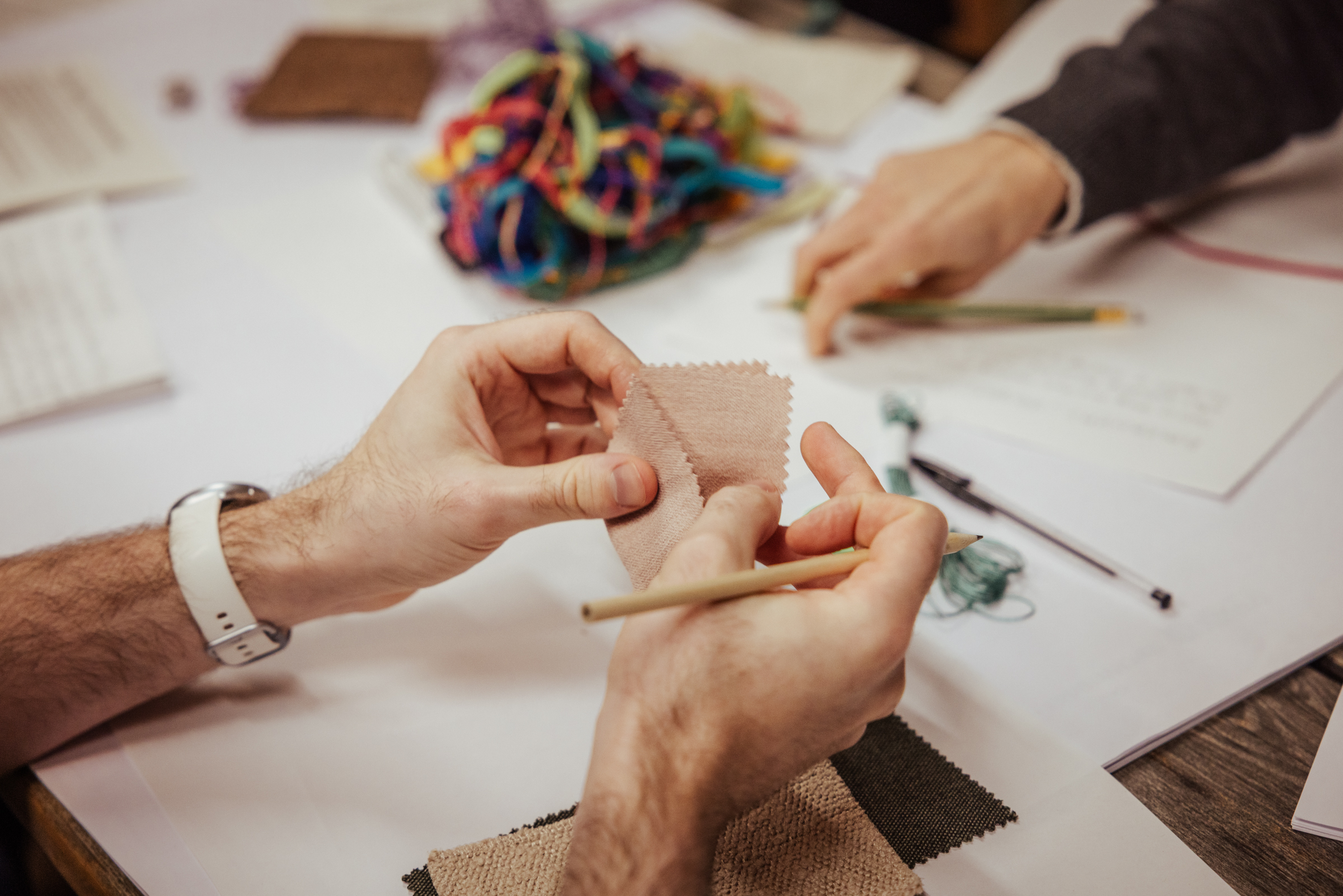 Closeup of workshop participants touching fabric samples during the workshop. While one person's two hands are holding and carefully caaressing a small piece of fabric, another person's hand is placing a pencil atop their writing. Various pieces of paper and more materials, such as strings, are scattered across the table.