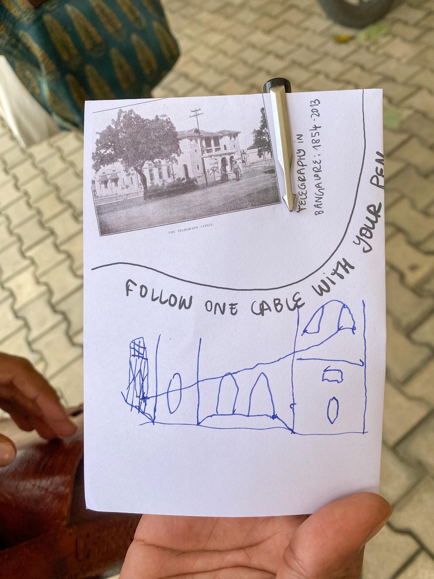 A person holds a sheet of paper from a zine that Iz has made for the network walk into the camera. They have drawn the Central Telegraph Office and the internet cables that span across it onto the front page with blue ink. Above their drawing there is a historic photo of the Central Telegraph Office printed, as well as the words ‚Follow one cable with your pen‘.