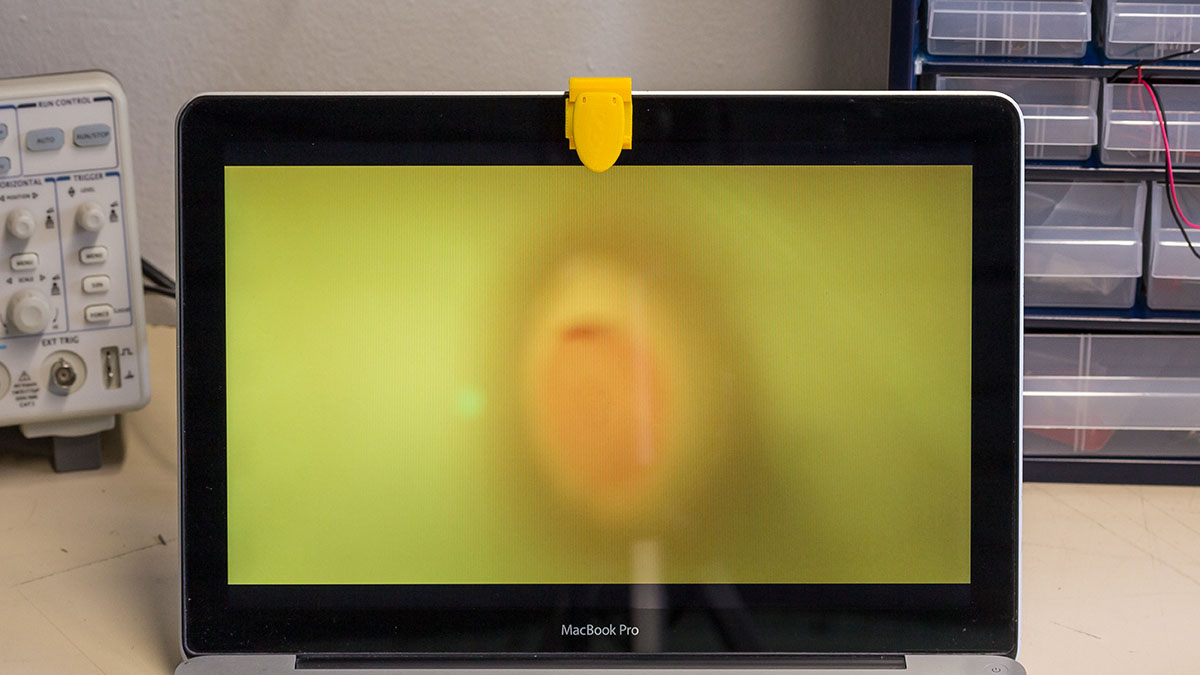 A macbook with the webcamera obscura in the shape of a toilet is positioned on a table amidst DIY hacking equipment. On the screen, one can see only yellow, since the lid of the toilet is closed.